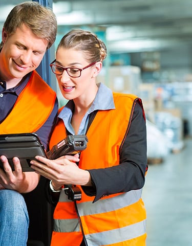 Gestion d'entrepôt : technicien et tablette Deux employés d'entrepôt souriants, portant des gilets haute visibilité, consultent ensemble une tablette et un scanner code-barres.