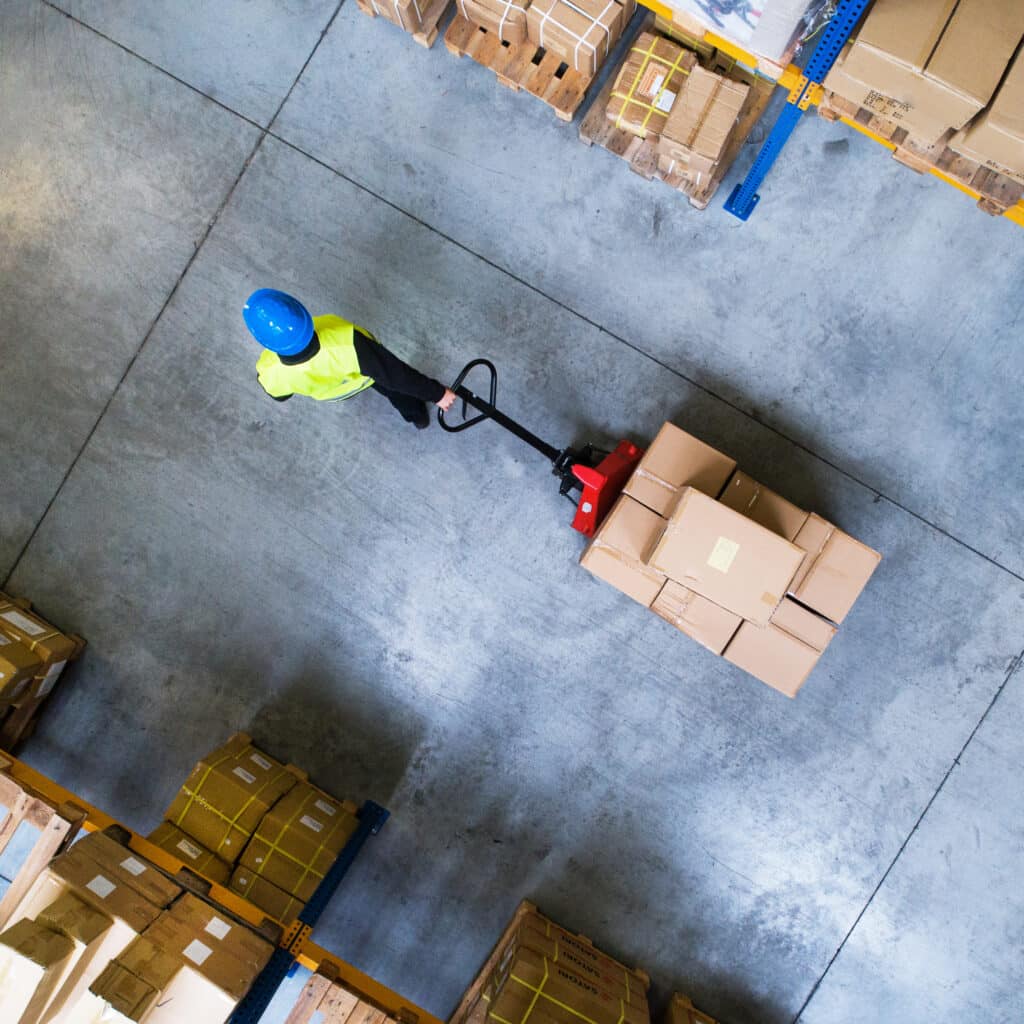 Entrepôt : Ouvrier déplace des cartons avec transpalette Vue plongeante d'un employé d'entrepôt avec casque bleu et gilet jaune déplaçant des cartons sur une palette avec un transpalette rouge.