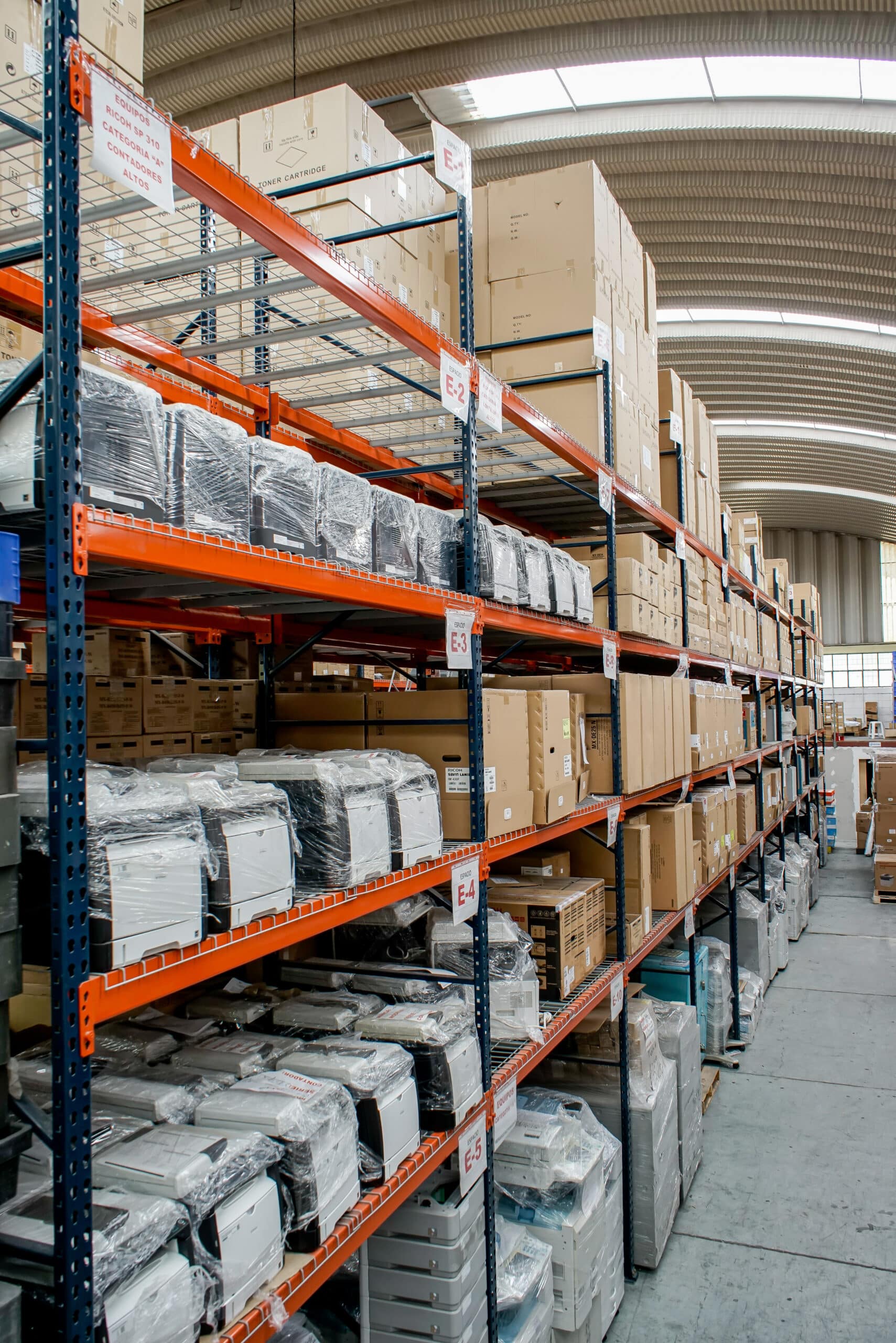 Intérieur d'un entrepôt avec rayonnages métalliques orange et bleus, stockant des imprimantes emballées et des cartons. Étiquettes visibles : "ESPACIO E-1" à "E-5", "EQUIPOS RICOH SP 310".