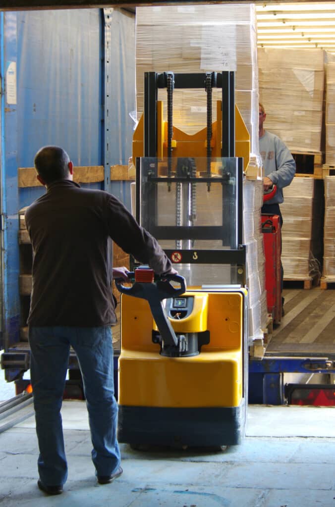 Manutentionnaire charge un camion avec transpalette électrique Un homme contrôle un transpalette électrique jaune devant une remorque de camion bleue chargée de palettes emballées sous film plastique.
